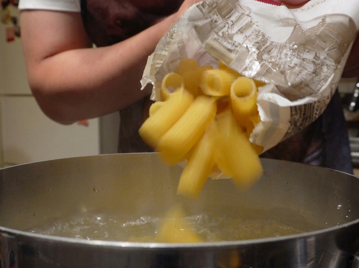 Dry rigatoni pasta falling from a paper bag into a pot of rapidly boiling water.