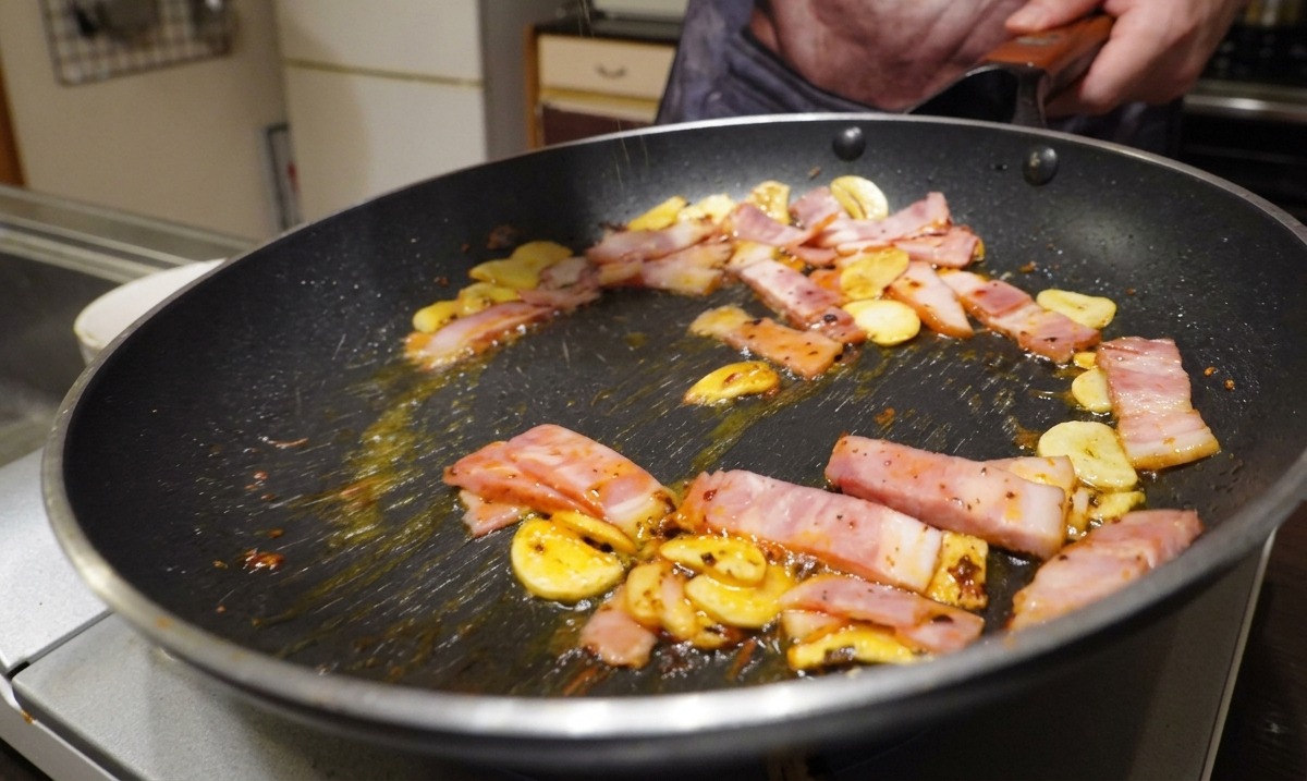 Sliced bacon, garlic, and chili flakes frying in a pan while being seasoned with freshly ground black pepper.