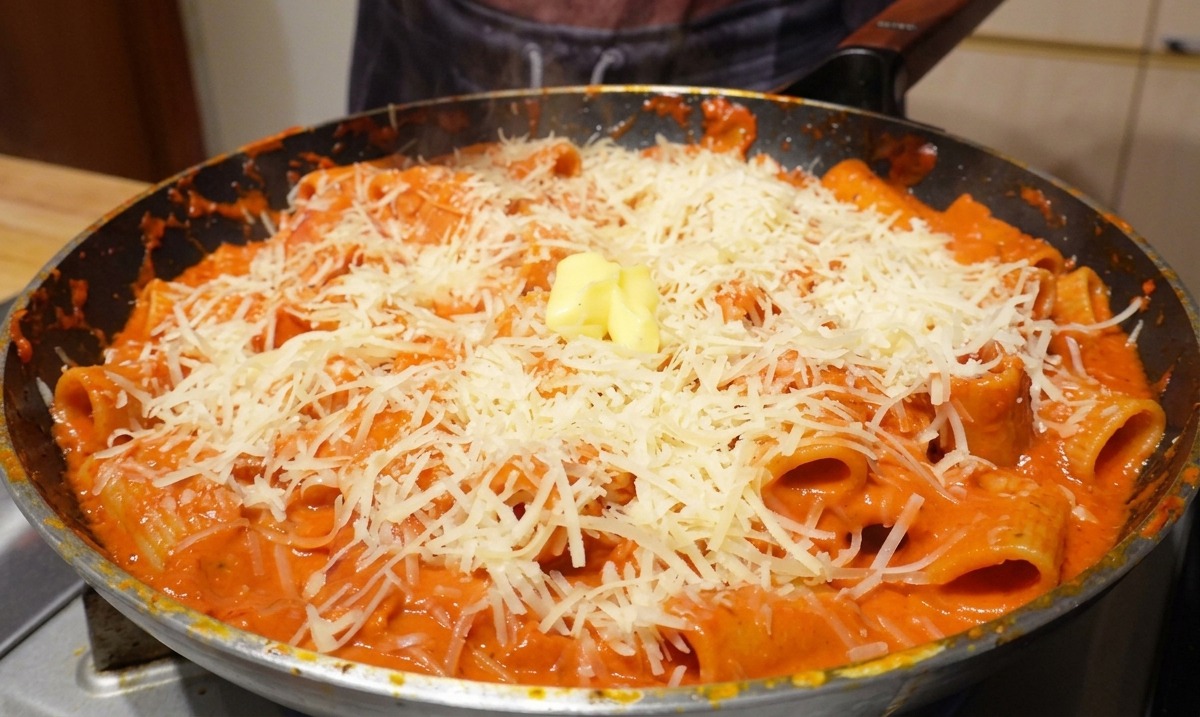 A small cube of yellow butter resting on top of a generous pile of grated parmesan and rigatoni pasta in a hot frying pan.