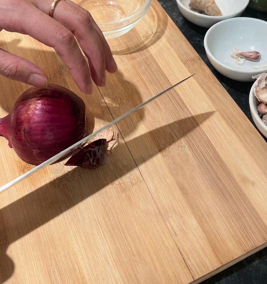 A hand holding a red onion on a wooden cutting board with a chefs knife resting against it, ready to slice.
