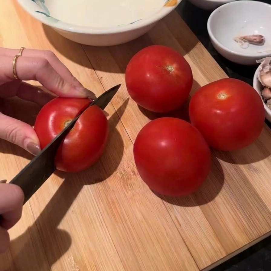 Hands using a chefs knife to slice a fresh red tomato on a wooden cutting board with three whole tomatoes nearby.