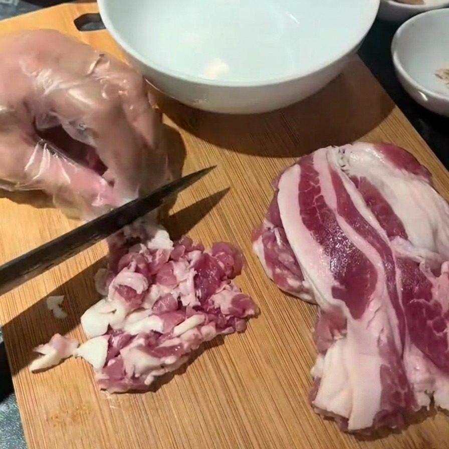 A gloved hand using a black knife to chop raw bacon strips into small pieces on a wooden cutting board.