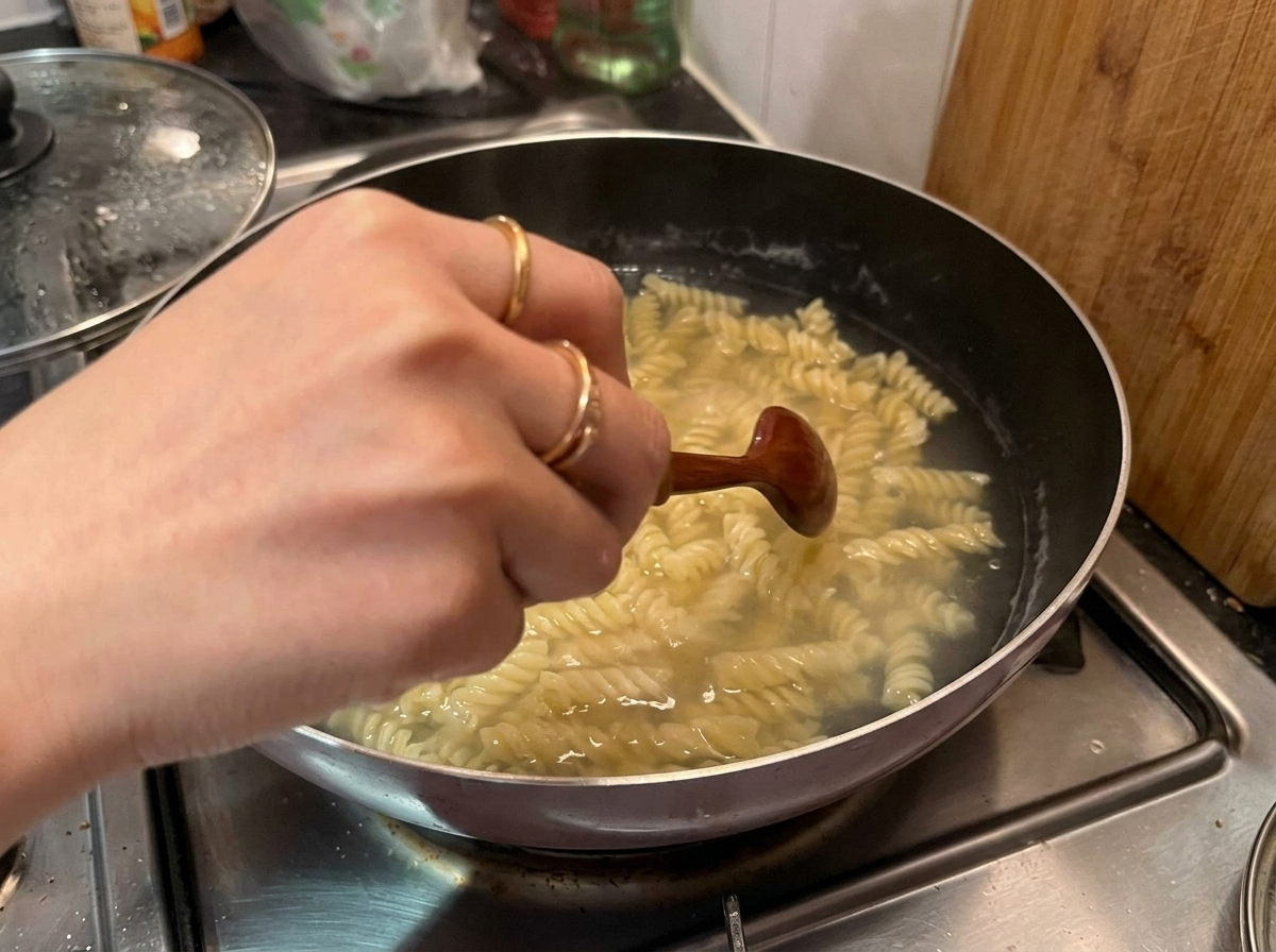 A hand using a small wooden spoon to stir and scoop cooked fusilli pasta out of a pot of boiling water.