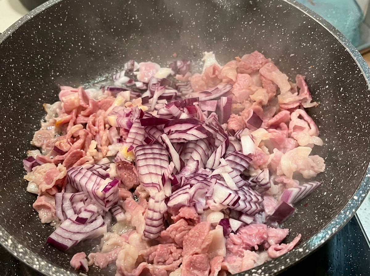 Diced red onion scattered over pieces of partially cooked bacon in a hot speckled skillet.