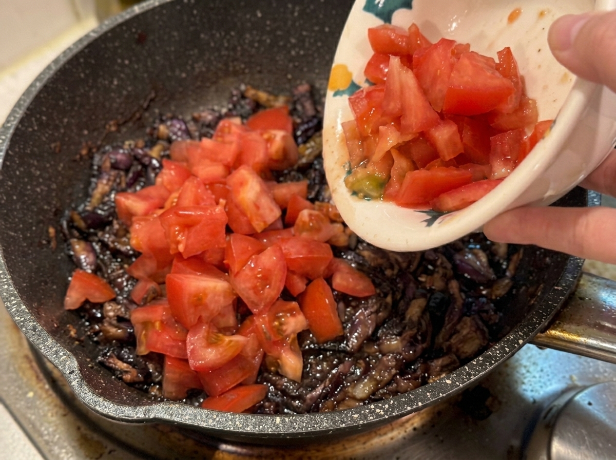 A hand holding a ceramic bowl to pour fresh diced tomatoes into a hot pan filled with cooked bacon and onions.
