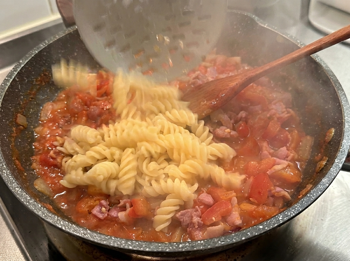 Cooked fusilli pasta being dropped from a slotted spoon into a hot pan of red tomato and bacon sauce.