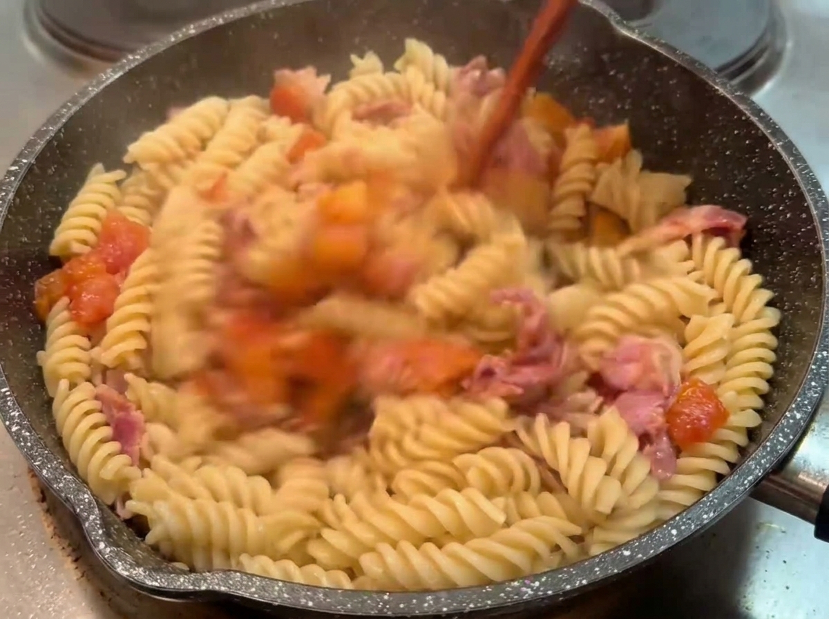 Cooked fusilli pasta being tossed in a dark pan with a chunky tomato and bacon sauce using a wooden utensil.