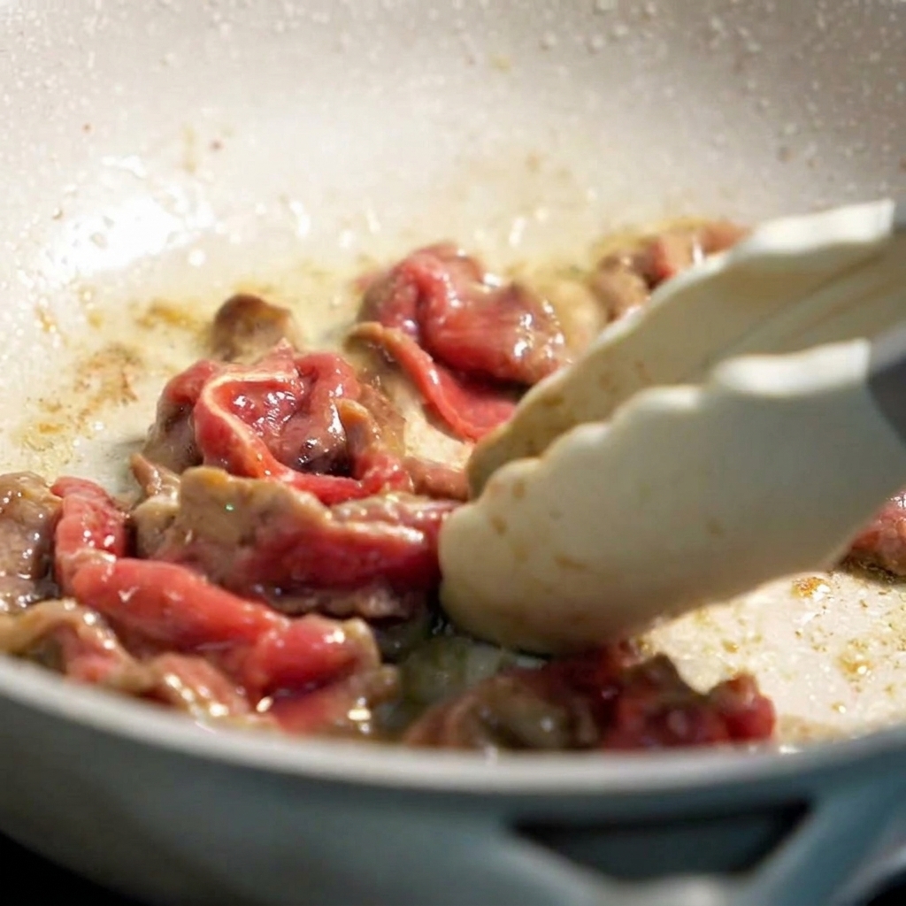Partially cooked thinly sliced beef being tossed in a lightly oiled pan with silicone tongs.