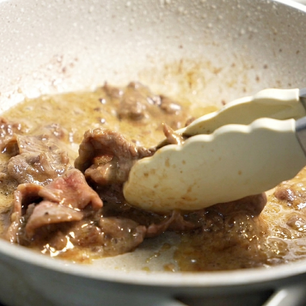Tongs tossing partially browned beef slices in a hot pan with cooking oil.