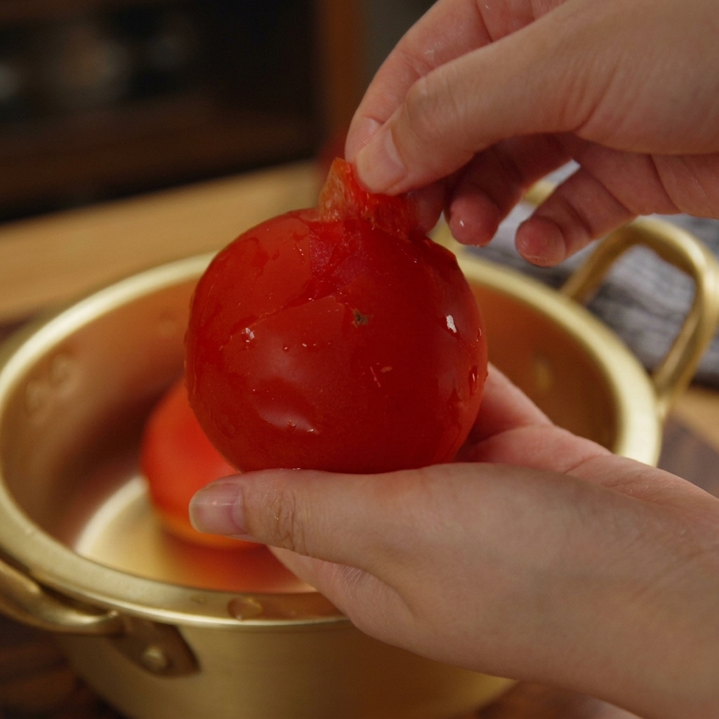 A persons hands peeling the loose skin off a bright red tomato over a gold bowl.