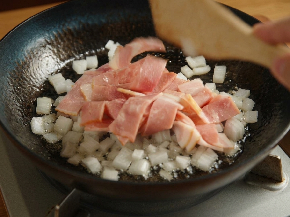 A wooden spatula mixing chopped bacon and diced white onions in a black skillet over a stovetop.