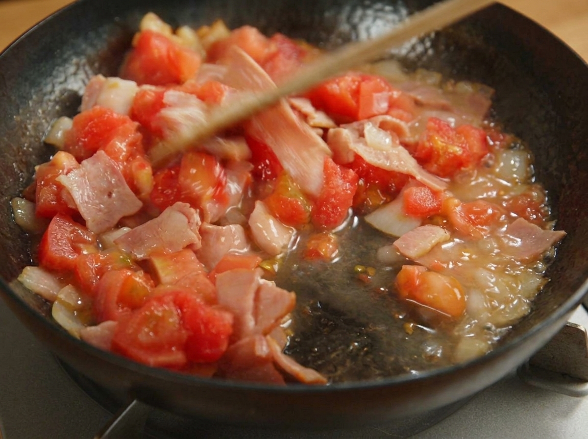 A wooden spatula stirring diced tomatoes, bacon, and onions in a black pan.