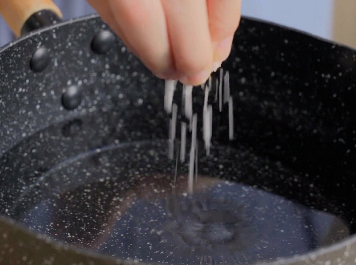 A hand sprinkling a pinch of coarse white salt into a dark pot filled with water.