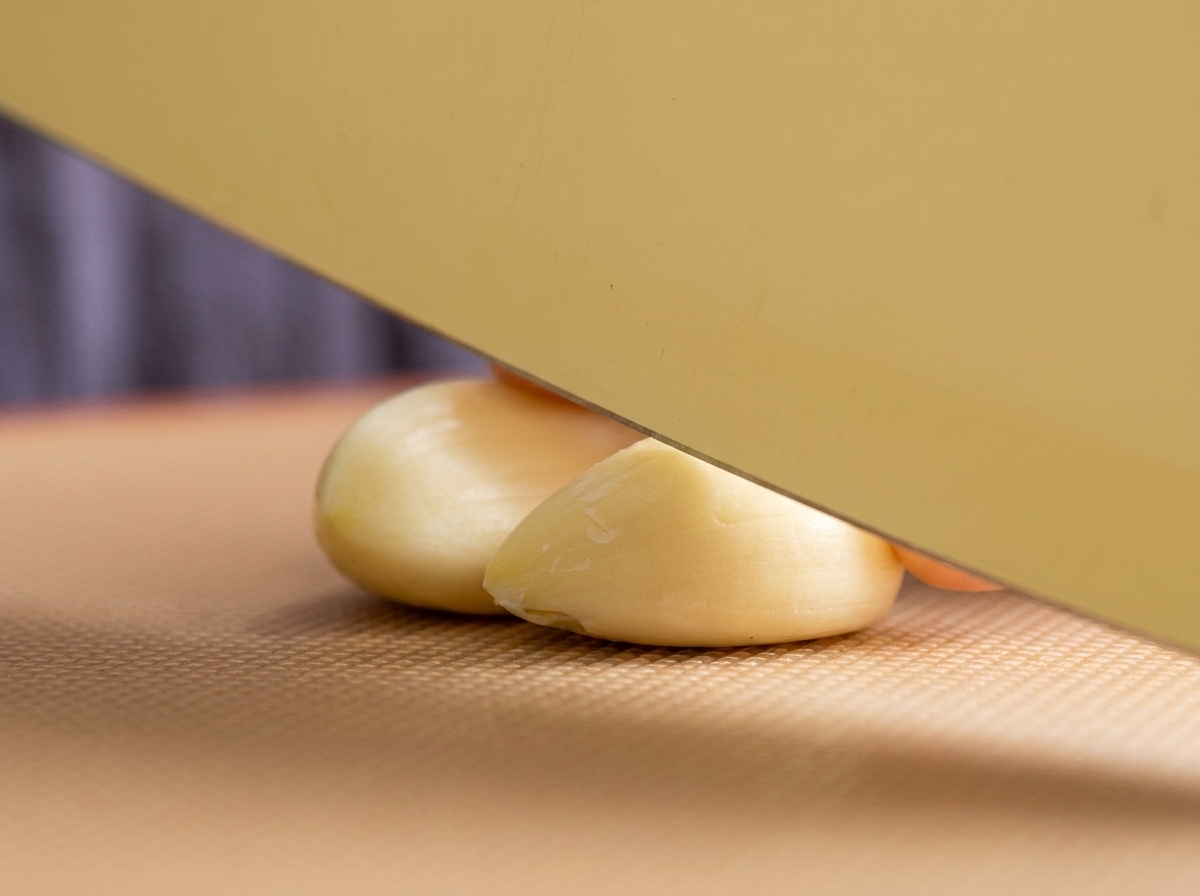 A wide kitchen knife resting on two peeled garlic cloves on a brown textured cutting board, ready to be chopped.