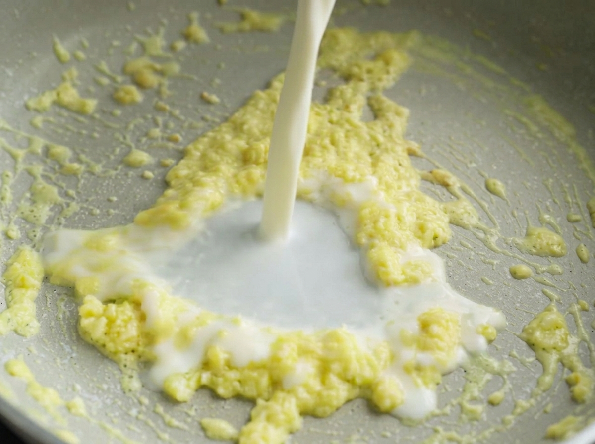 A stream of white milk being poured into a pan containing a sizzling yellow butter and minced garlic mixture.
