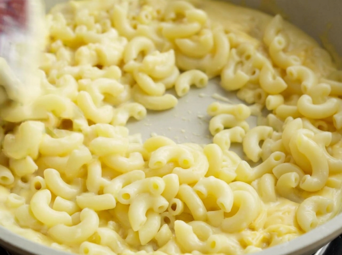 A spatula tossing cooked macaroni in a light-colored pan, thoroughly coating the pasta in creamy cheese sauce.