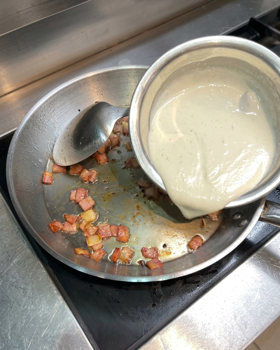 Liquid cream being poured from a small bowl into a pan containing crispy bacon.