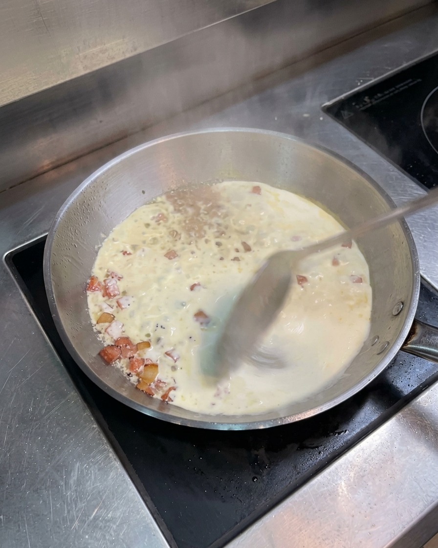 A stainless steel pan filled with simmering cream and crispy bacon bits, being stirred with a metal spoon.