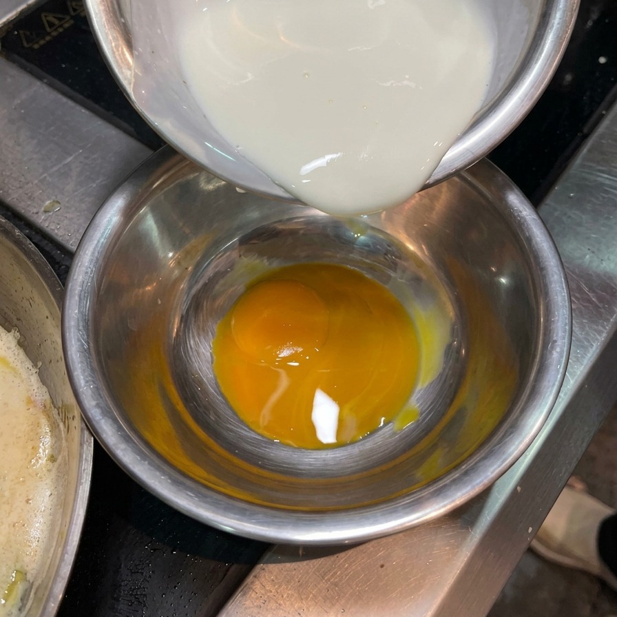 Liquid cream being poured from a small bowl into another bowl containing a bright orange raw egg yolk.