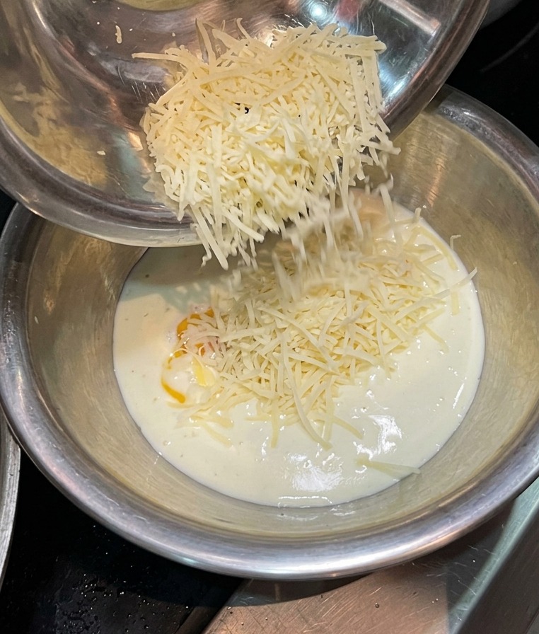 Grated cheese falling from a metal bowl into a mixture of liquid cream and egg yolk.