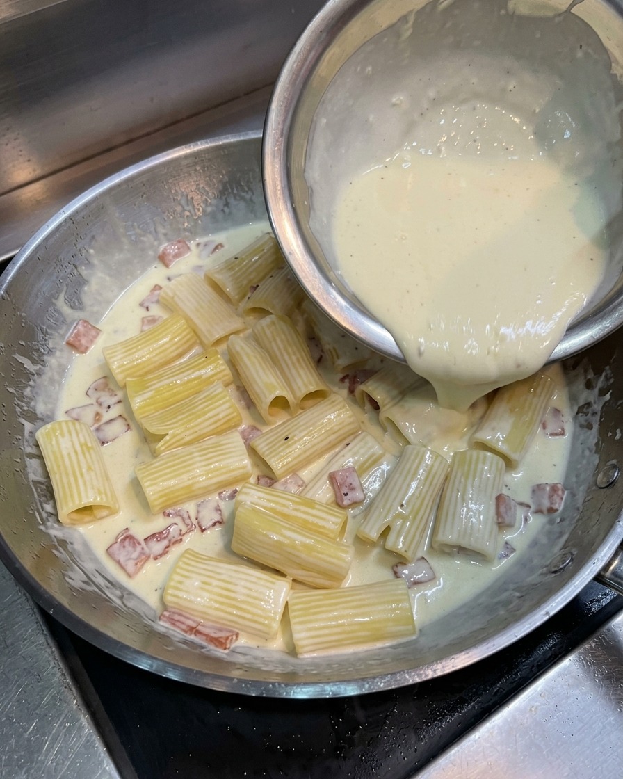 Pouring a pale yellow mixture of egg yolk, cream, and cheese from a metal bowl over rigatoni pasta and bacon in a skillet.