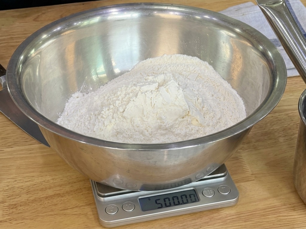 A large stainless steel mixing bowl on a digital scale containing a mound of white high-gluten flour.