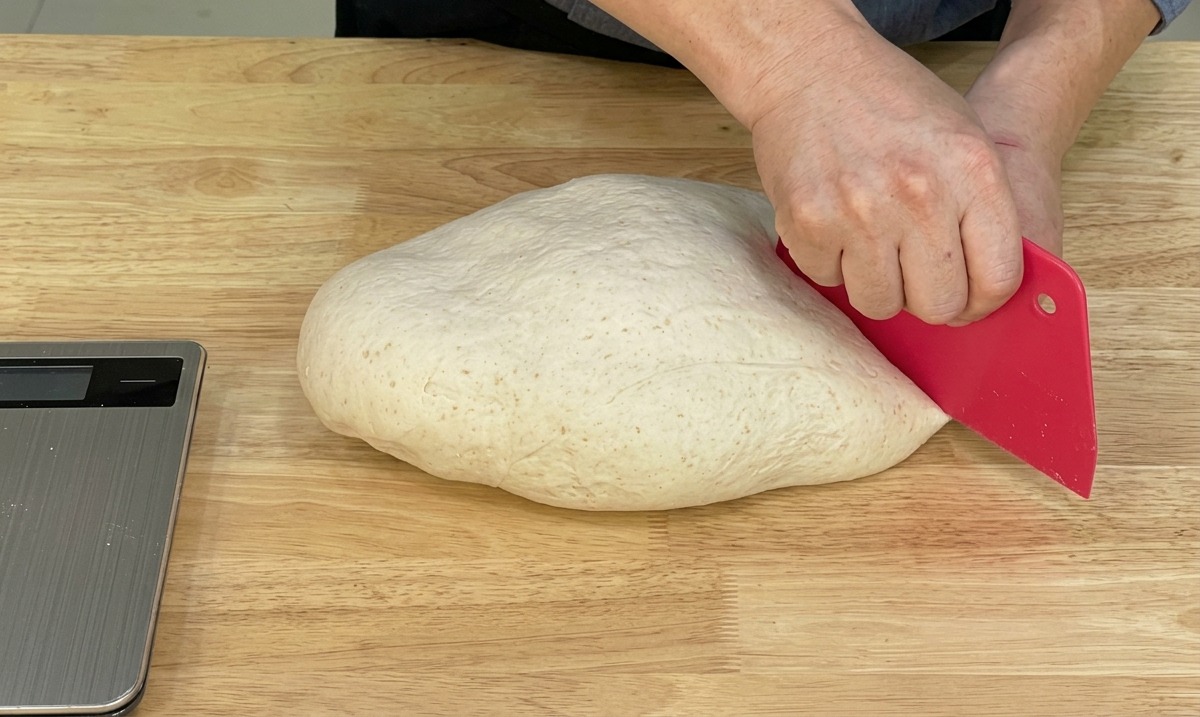 Chef using a red bench scraper to divide a large piece of smooth pizza dough on a wooden surface.