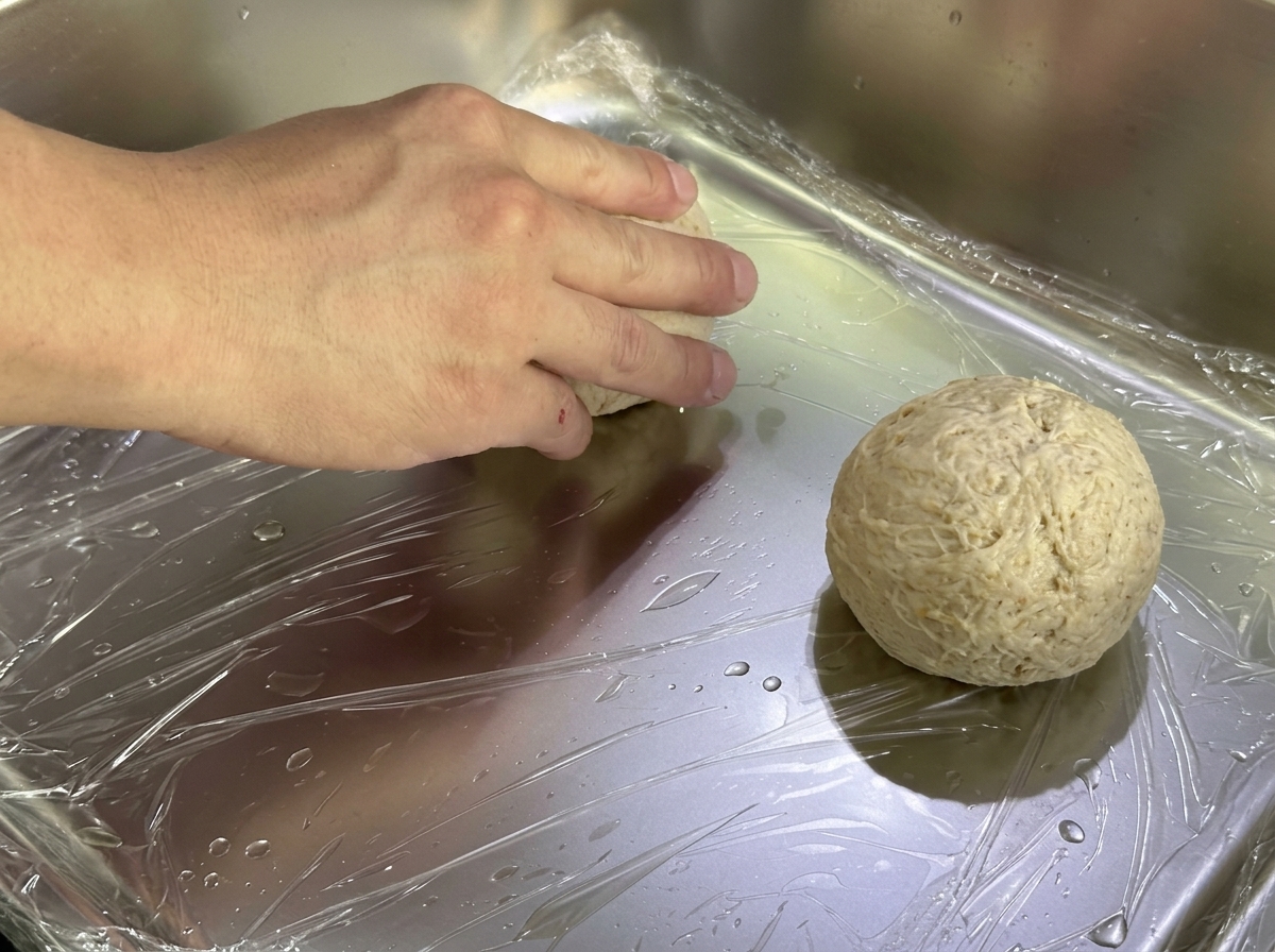 Placing shaped dough balls onto a metal tray covered with plastic wrap for proofing.