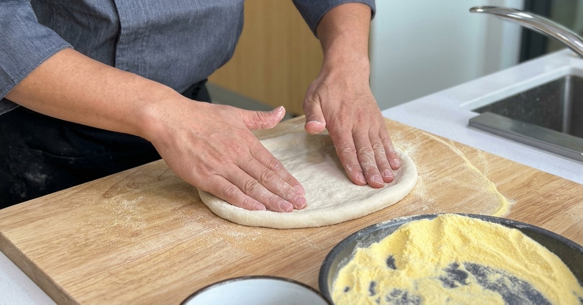 Stretching the pizza dough base outward on a wooden board, maintaining a thicker raised edge.