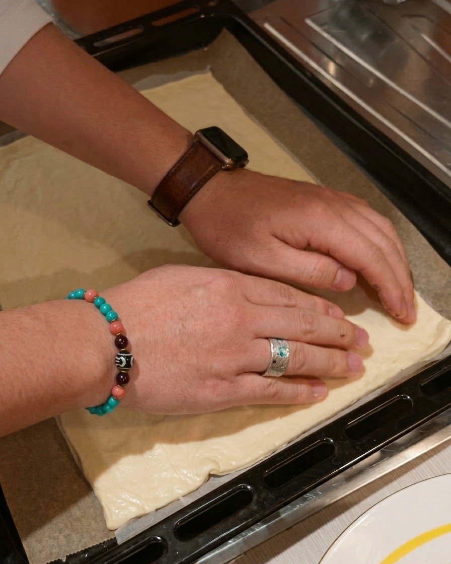 Two hands gently pressing down on the edges of a rectangular sheet of raw pizza dough resting on a dark baking tray.
