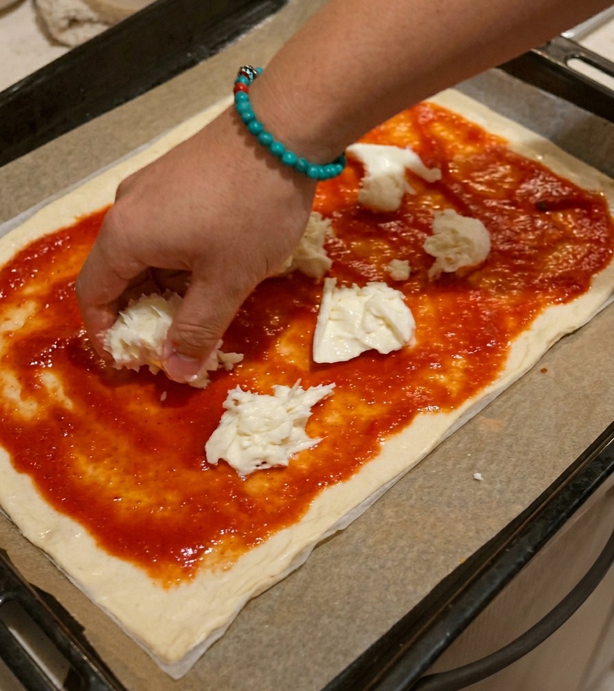A hand placing unevenly torn chunks of white mozzarella cheese onto a pizza crust coated with red tomato sauce.