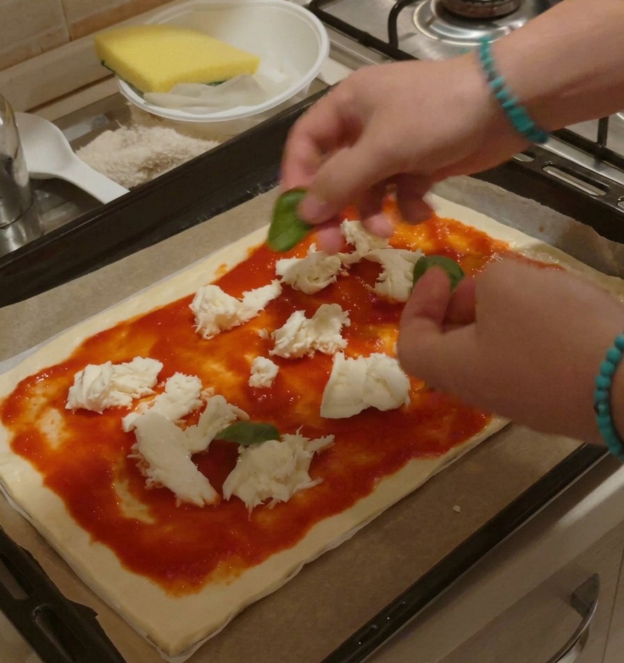 Hands carefully arranging whole, fresh green basil leaves on an unbaked pizza covered with tomato sauce and scattered mozzarella chunks.