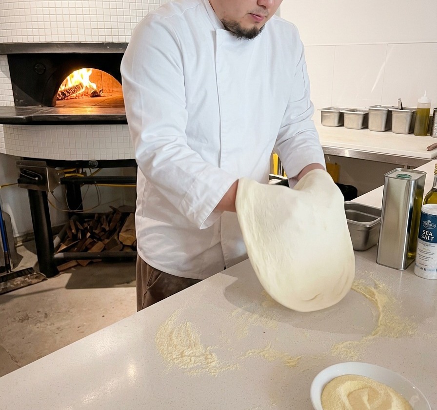 A chef pressing and stretching a round pizza dough on a white countertop dusted with flour.
