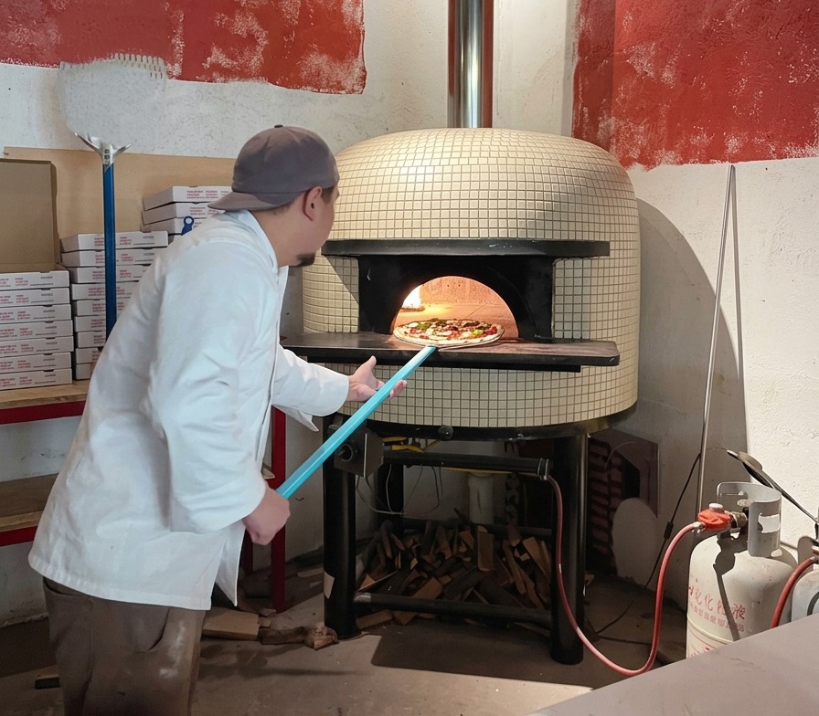 A chef using a long blue pizza peel to slide an uncooked pizza into the mouth of a hot wood-fired brick oven.