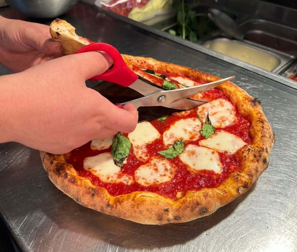 Hands using red-handled kitchen scissors to cut a hot, bubbly Margherita pizza on a stainless steel counter.