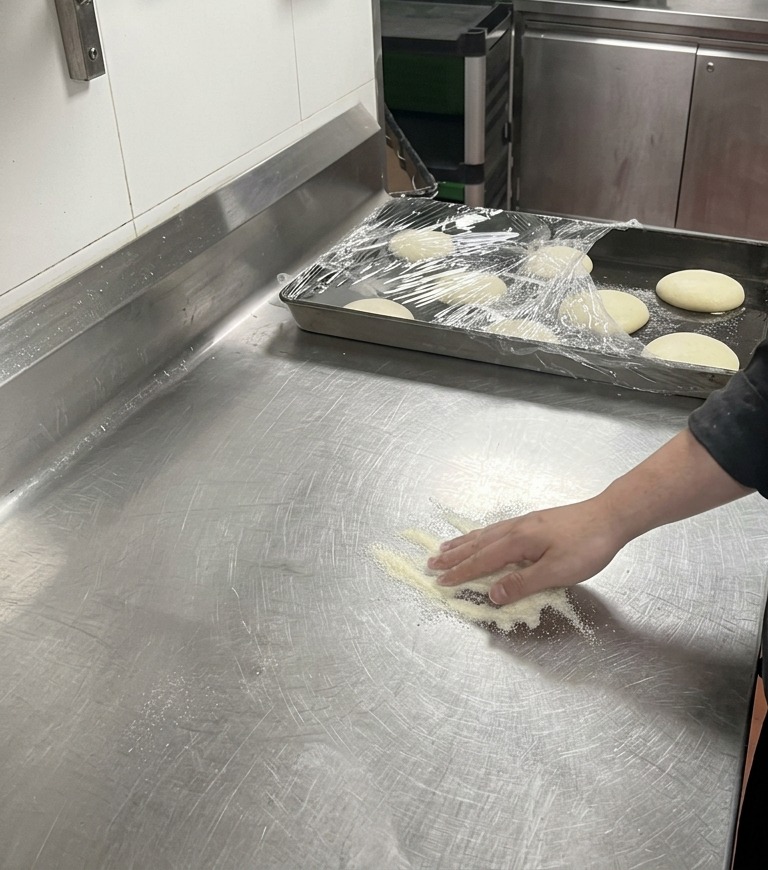 A chefs hand gently pressing a ball of pizza dough into a pile of white dusting flour on a metal countertop.