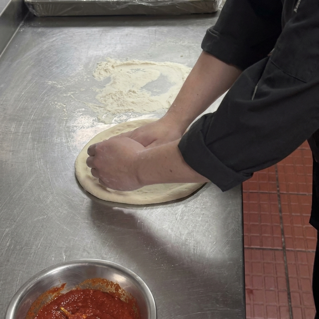 Hands pressing down the center of a flattened pizza dough, leaving a thick rim around the edge.