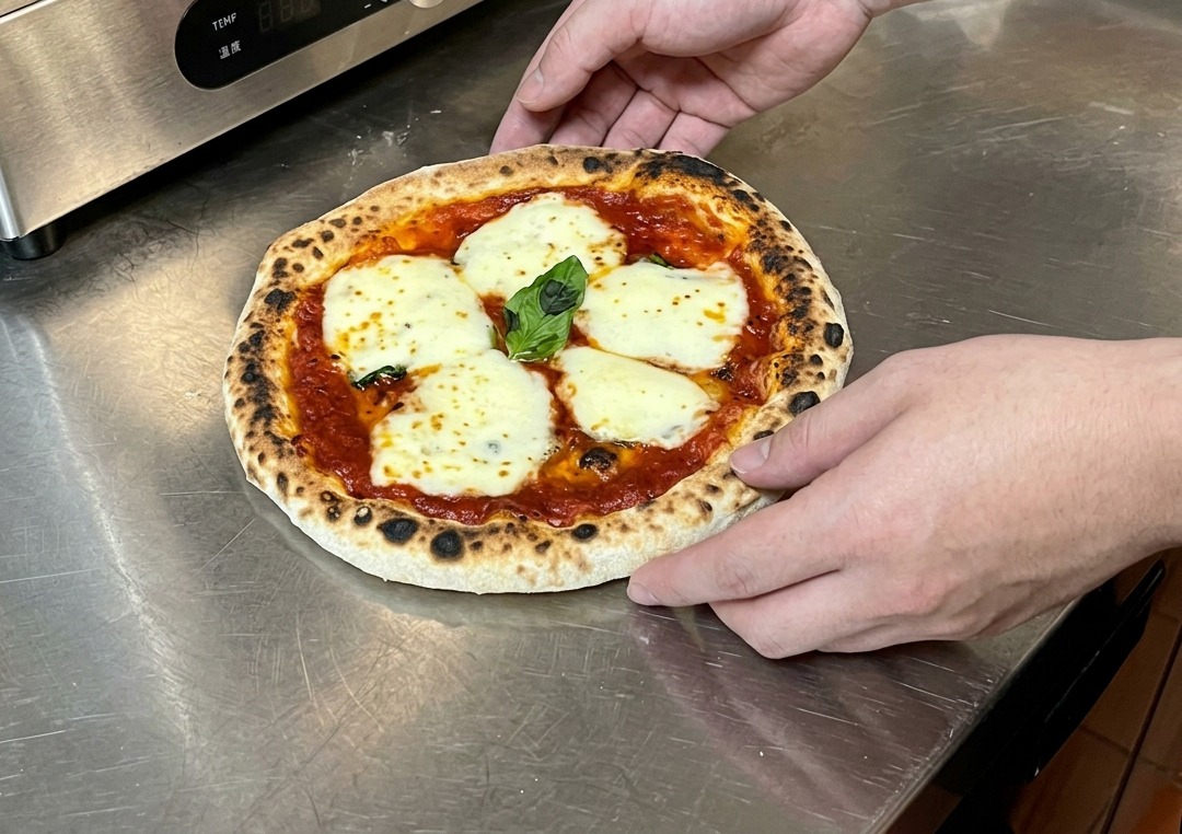 Hands holding a freshly baked pizza on a metal countertop, preparing to check the crispness of the bottom crust.