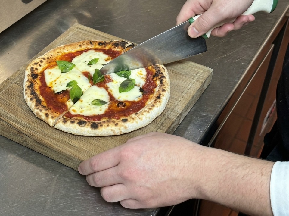 Hands using a large knife to slice a fresh Margherita pizza into wedges on a wooden cutting board.