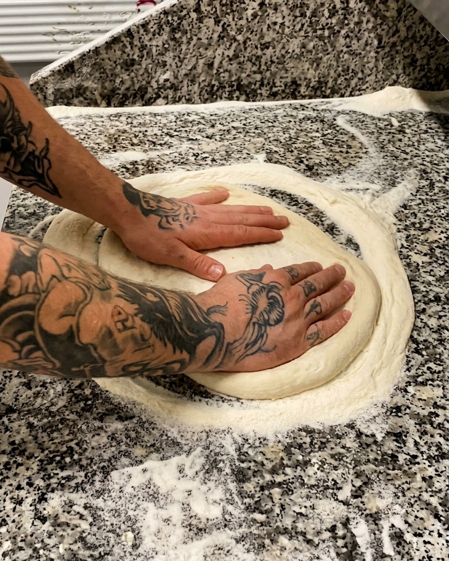 Tattooed hands gently pressing and stretching a round piece of pizza dough on a floured marble surface.