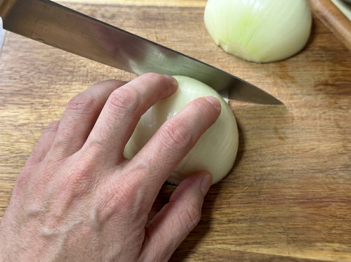 A hand slicing a peeled white onion into thin strips on a wooden cutting board.