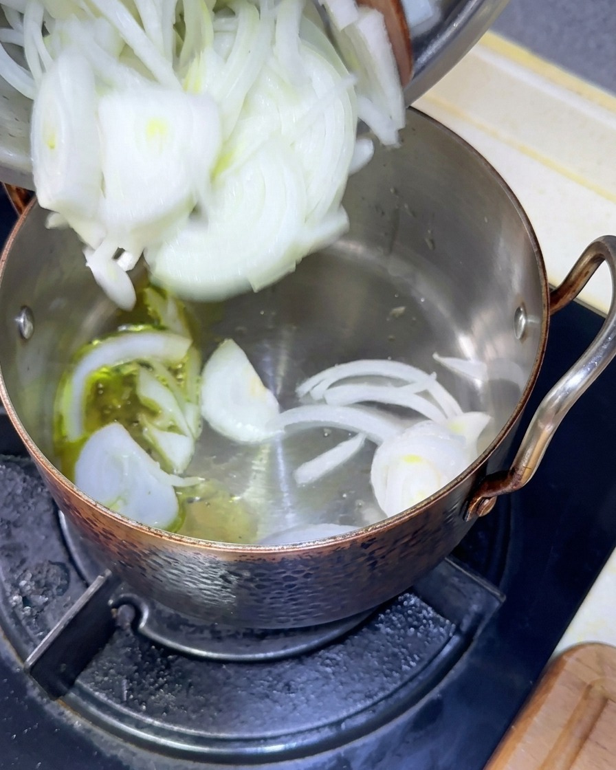 Freshly sliced white onions being poured from a bowl into a copper pot with warm olive oil.