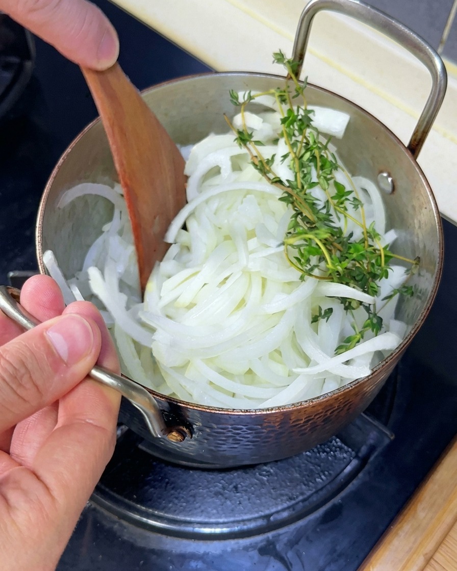 Sliced white onions and fresh sprigs of thyme being stirred with a wooden spatula in a copper pot.