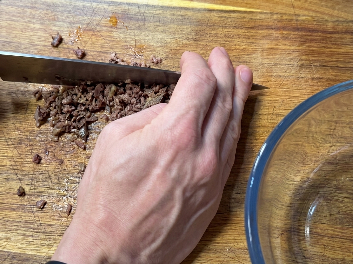 A hand using a large knife to finely mince anchovy fillets on a wooden cutting board.