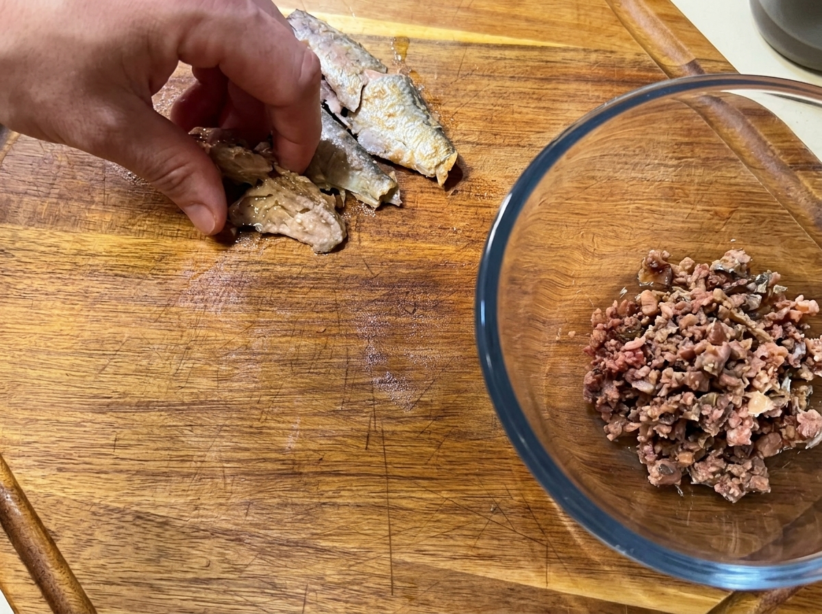 A hand breaking apart deboned canned sardine fillets on a cutting board, next to a bowl of minced fish.