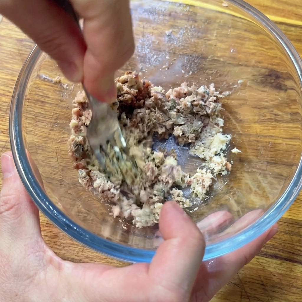 Hands using a metal fork to mash minced anchovies and sardines into a paste inside a clear glass bowl.
