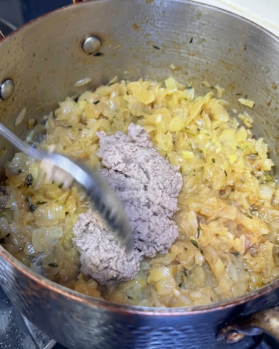 A spoonful of greyish mashed fish paste being added into a pot of deeply caramelized onions.