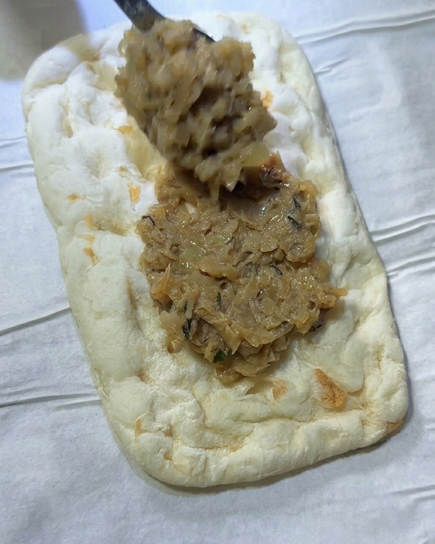 A spoonful of savory onion mixture being spread evenly across a rectangular piece of flatbread dough.