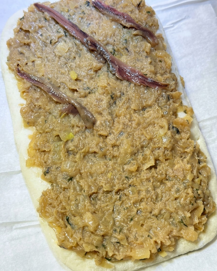 Anchovy fillets being arranged in a criss-cross diamond pattern on top of a thick layer of onion paste spread over flatbread dough.