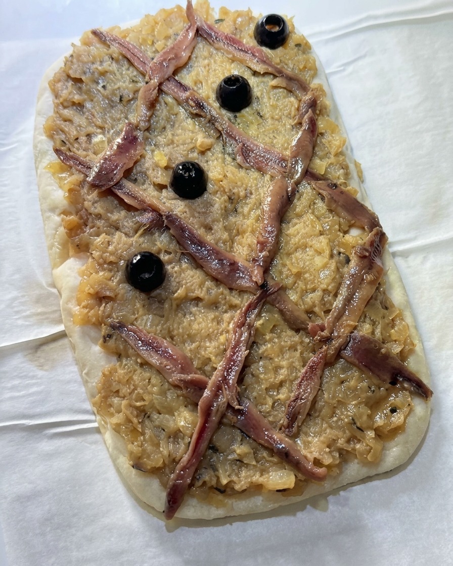 Whole black olives placed into the empty diamond-shaped spaces of an anchovy lattice on top of an onion-covered flatbread.